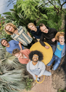 Seven people stand and sit around a small round yellow table in an outdoor setting with lush green plants, viewed from above.
